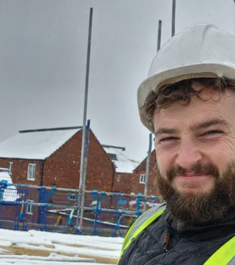 Ben on a snow covered housing development, in a hard hat and a hi-vis vest over a work gilet, with new houses and scaffolding behind him.