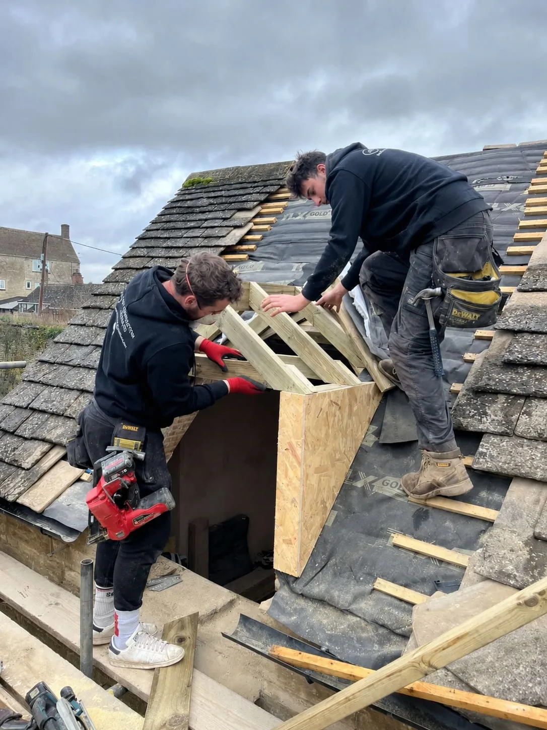 Two men on a stone slate roof, framing a dormer opening in timber, with the village rooftops behind them.