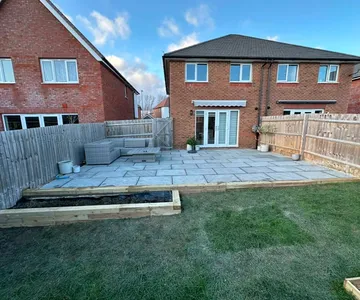 A patio laid in Indian sandstone across the back of a house, with a sleeper flowerbed built along the edge where it meets the lawn.