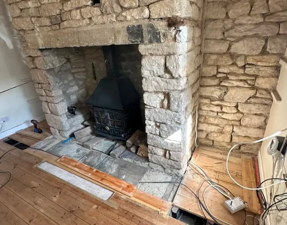 A stone inglenook with a cast iron stove in the opening, the chimney breast back to bare stone and the floor up in front of it.